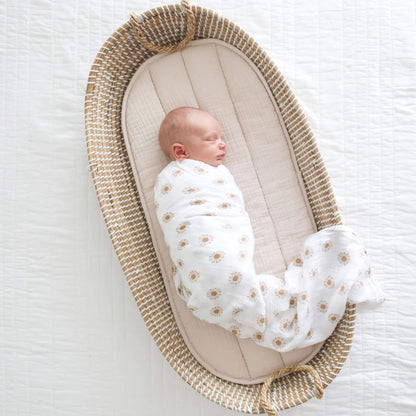 Baby resting in a woven basket, wrapped in sun-printed muslin swaddle.