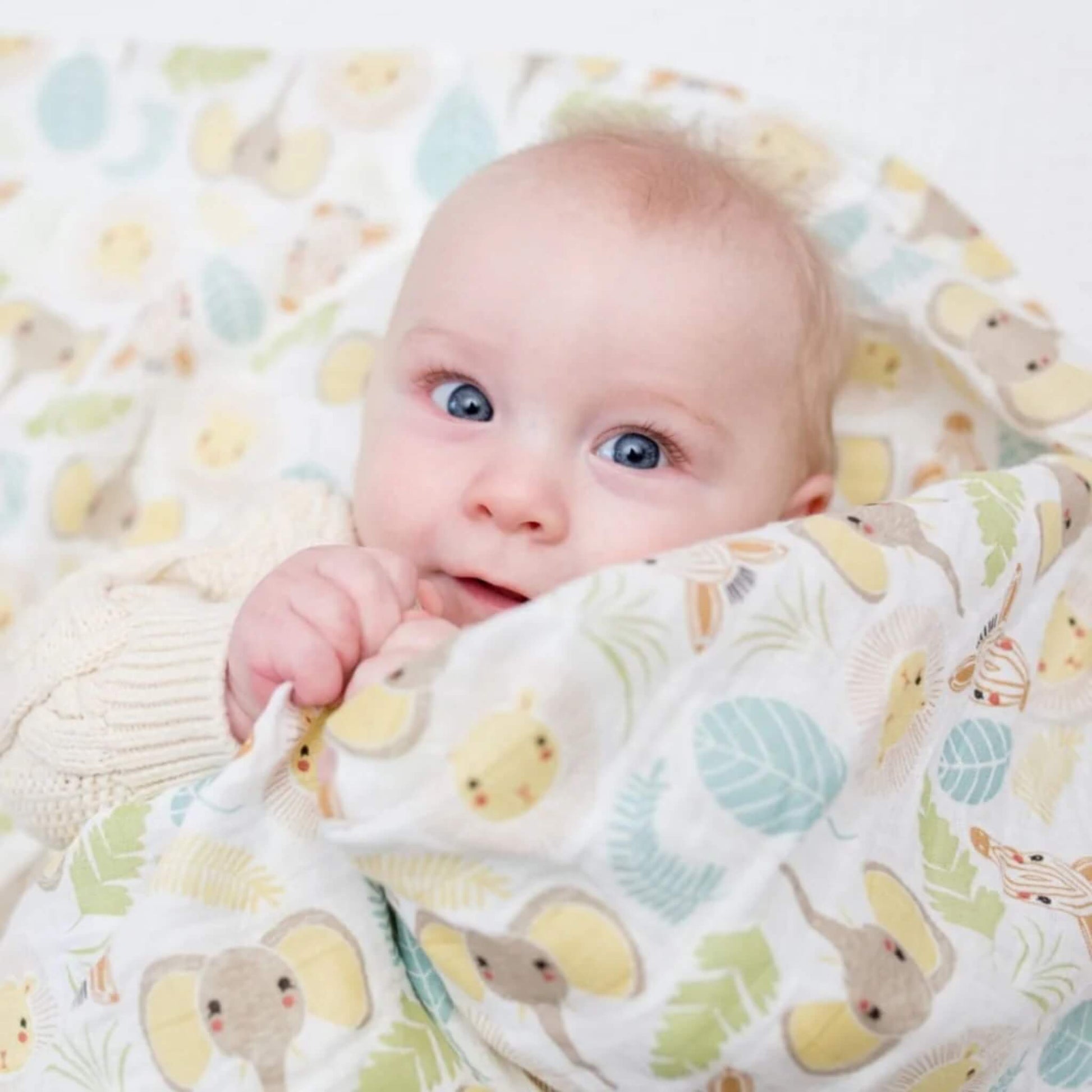 Baby wrapped in the Jungle Safari muslin swaddle, smiling while snuggled in the soft cotton fabric patterned with gentle safari animals and leaves.