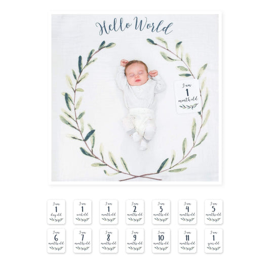 Baby lying on Lulujo Hello World milestone blanket with botanical wreath design showing 1 month milestone card, alongside complete set of age cards from 1 day to 1 year.
