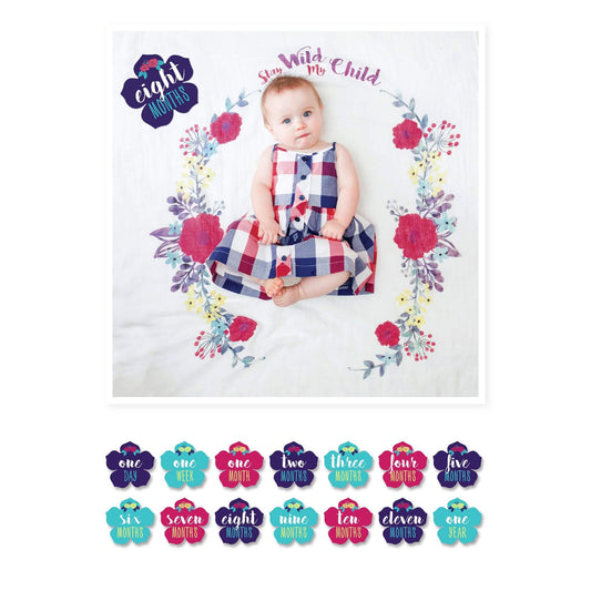 Lulujo "Stay Wild My Child" Milestone Muslin Blanket featuring 8-month-old baby girl in plaid dress with vibrant floral wreath design and flower-shaped milestone card.