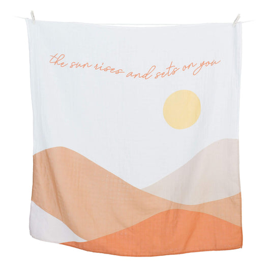 Lulujo white muslin blanket with 'The sun rises and sets on you' quote, yellow sun and warm earth tone waves design hanging on clothesline.