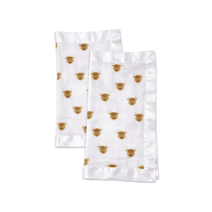A pair of folded muslin security blankets in white fabric with brown highland cow print and smooth satin borders.