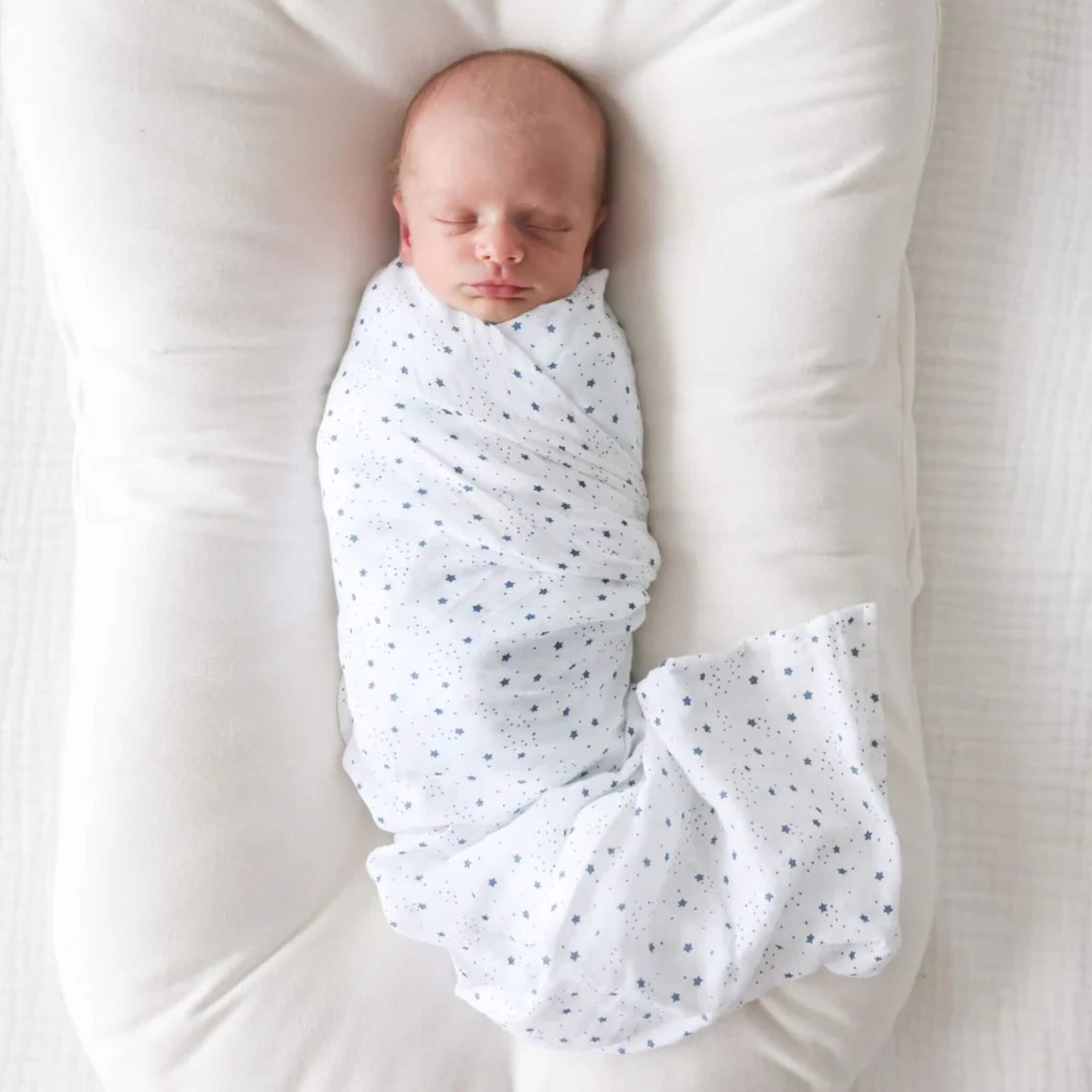 Overhead view of baby lying on a white nest, swaddled in a star-patterned muslin blanket.