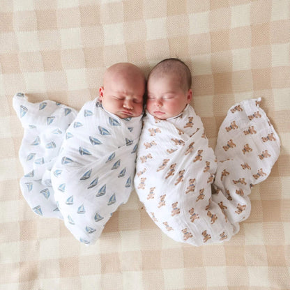Two newborn babies lying on a neutral blanket, each wrapped in a white muslin swaddle — one with a teddy bear print, the other with blue sailboats.