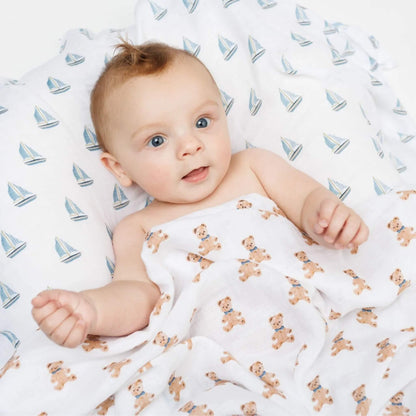 Baby lying under white muslin swaddles printed with teddy bears and blue sailboats, looking up with a happy expression