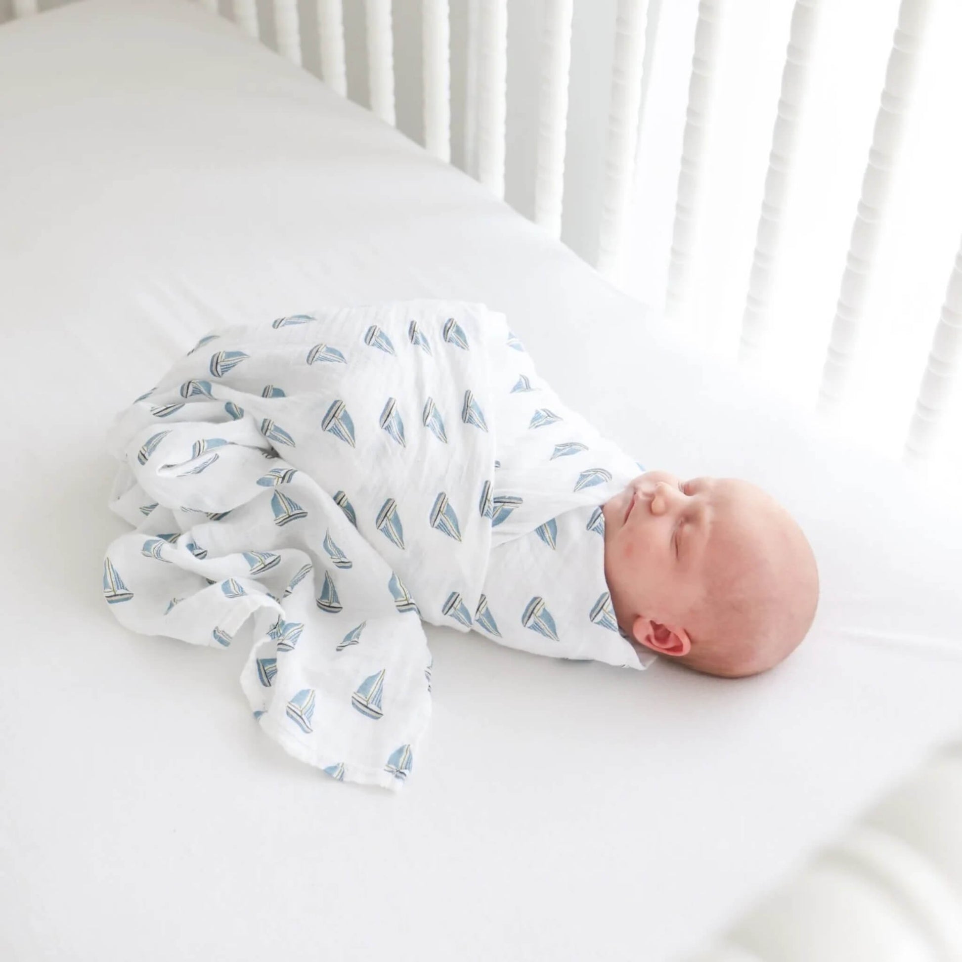 Sleeping baby swaddled in a white muslin blanket decorated with small blue sailboats.