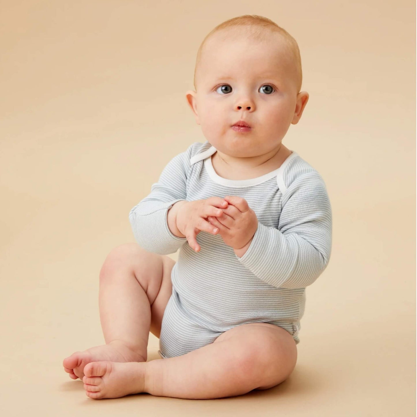 A baby sitting on a neutral backdrop wearing a blue striped long sleeve bodysuit, with hands together and looking slightly upward.