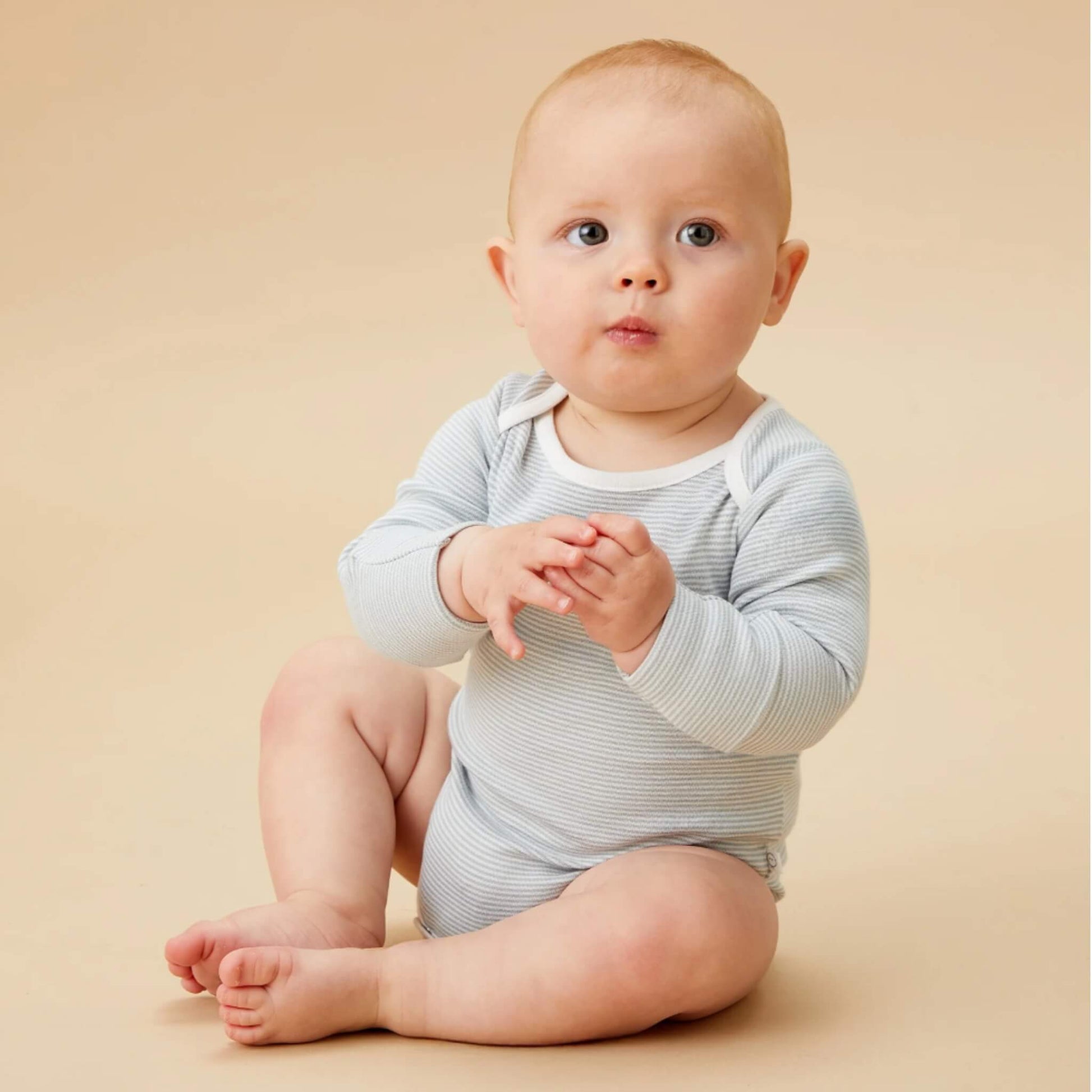 A baby sitting on a neutral backdrop wearing a blue striped long sleeve bodysuit, with hands together and looking slightly upward.