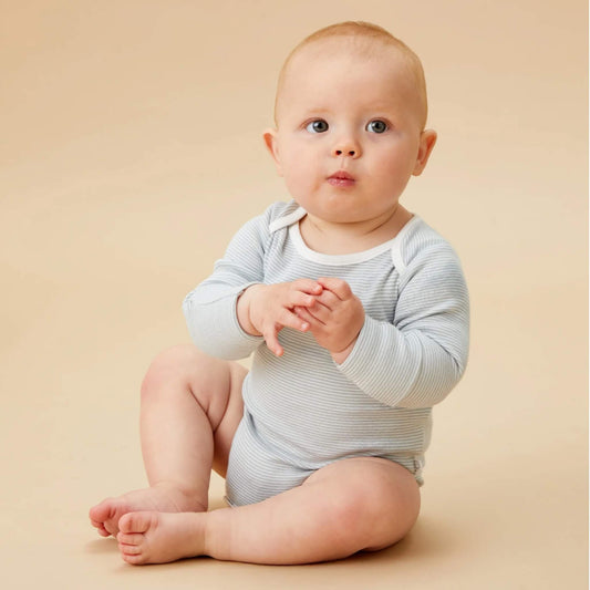 A baby sitting on a neutral backdrop wearing a blue striped long sleeve bodysuit, with hands together and looking slightly upward.