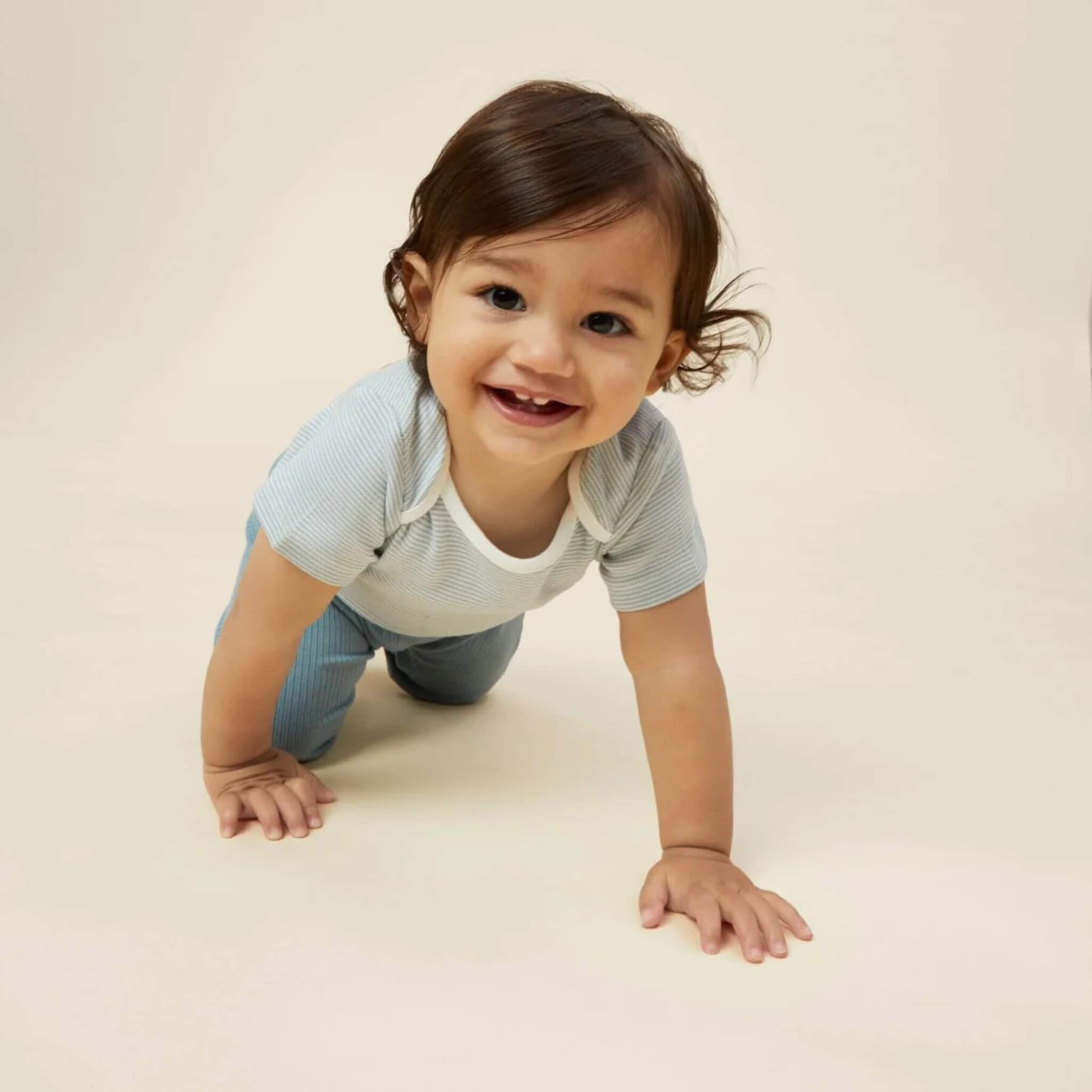 A baby crawling on a light surface while wearing a blue striped short sleeve bodysuit, smiling towards the camera.