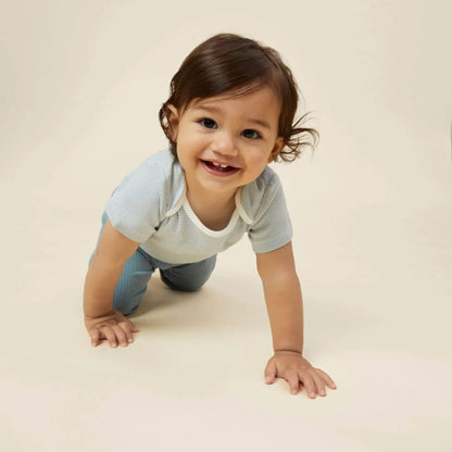 A baby crawling on a light surface while wearing a blue striped short sleeve bodysuit, smiling towards the camera.