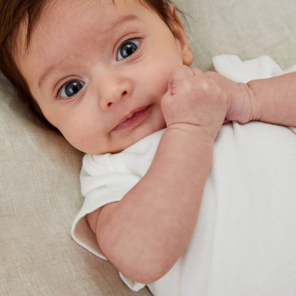 A young baby lying on a cushion wearing a short-sleeved white bodysuit, with soft natural light highlighting the fabric and their facial features.