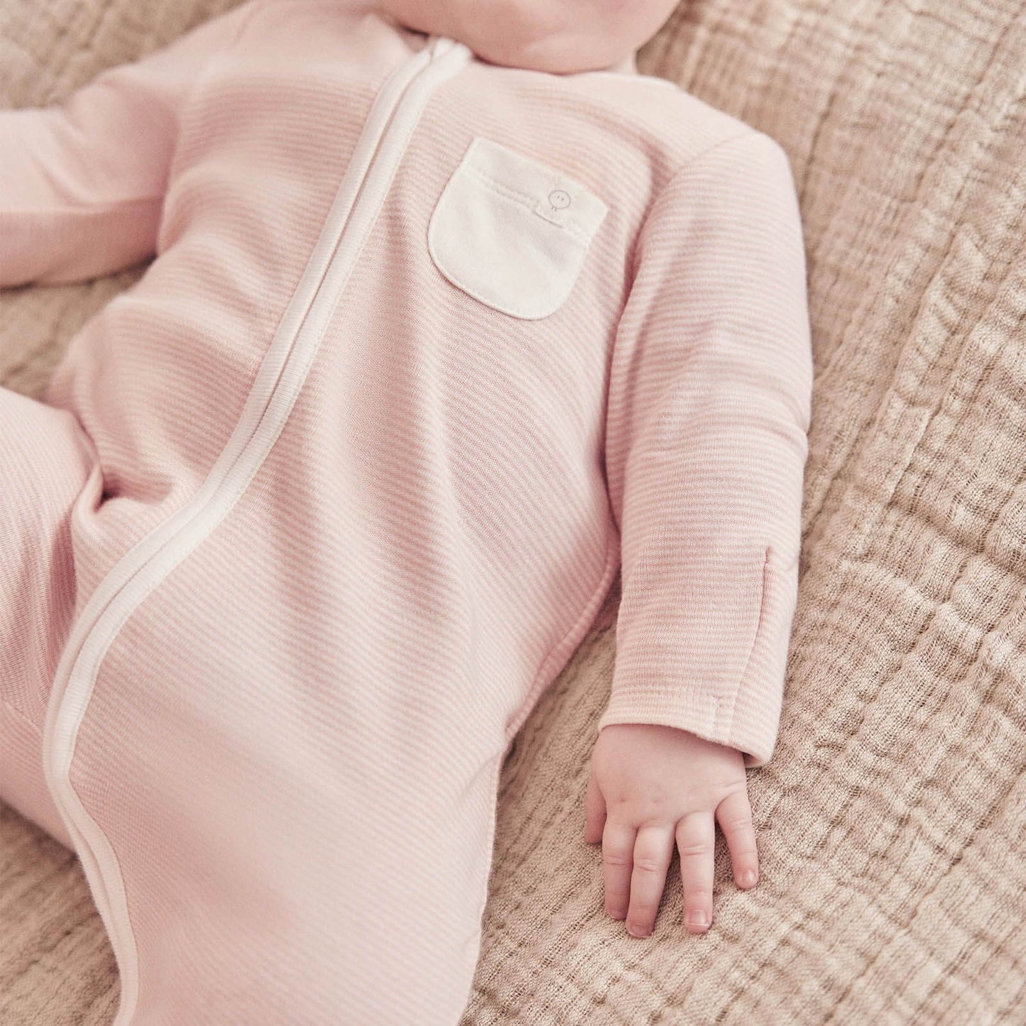 A baby lying on a textured blanket wearing a pink striped sleepsuit with a long front zip and built-in feet.