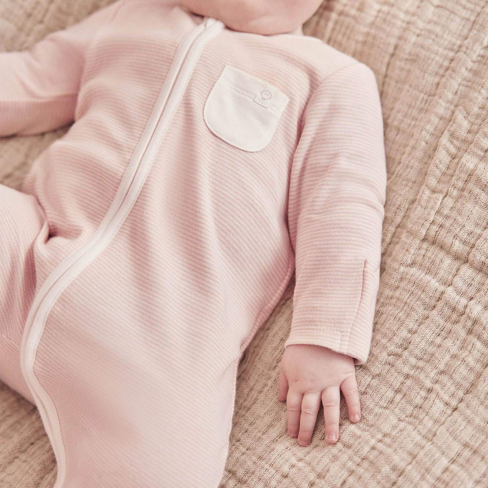 A baby lying on a textured blanket wearing a pink striped sleepsuit with a long front zip and built-in feet.