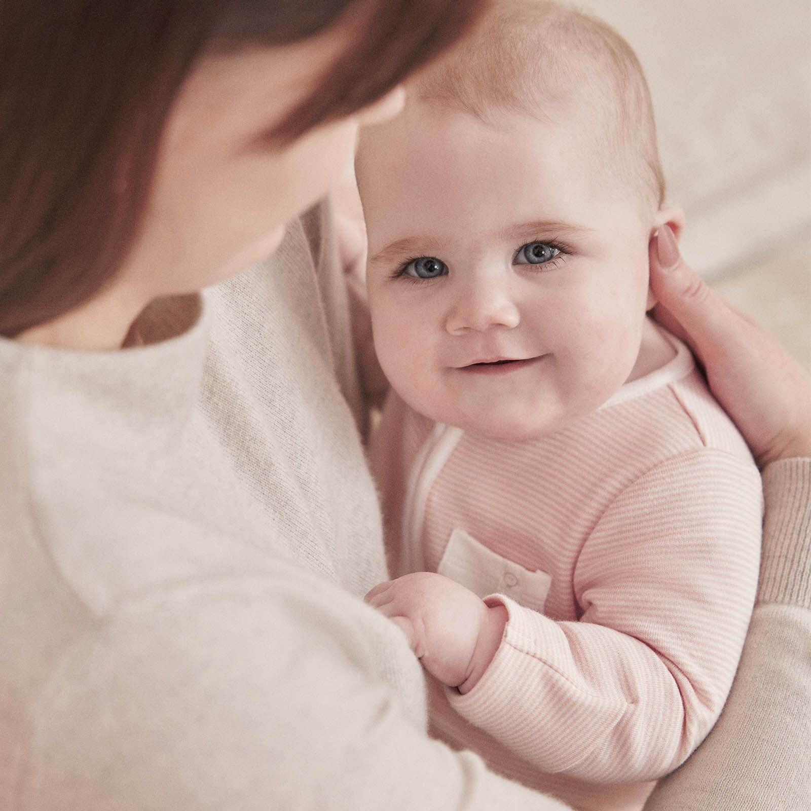 A baby wearing a pink striped sleepsuit resting in an adult’s arms, looking up and smiling.