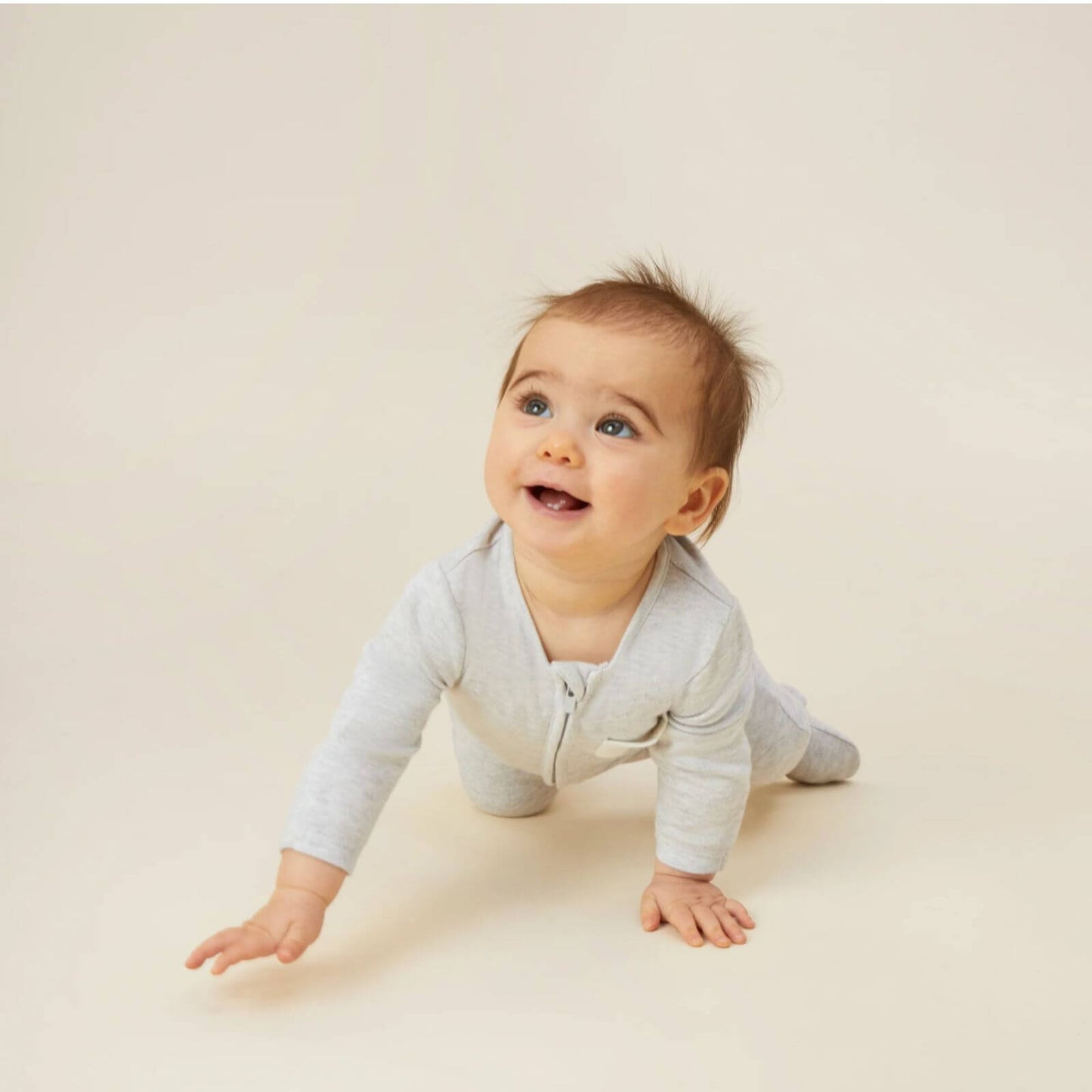 A baby crawling on a light surface while wearing a grey sleepsuit with a long front zip and long sleeves.