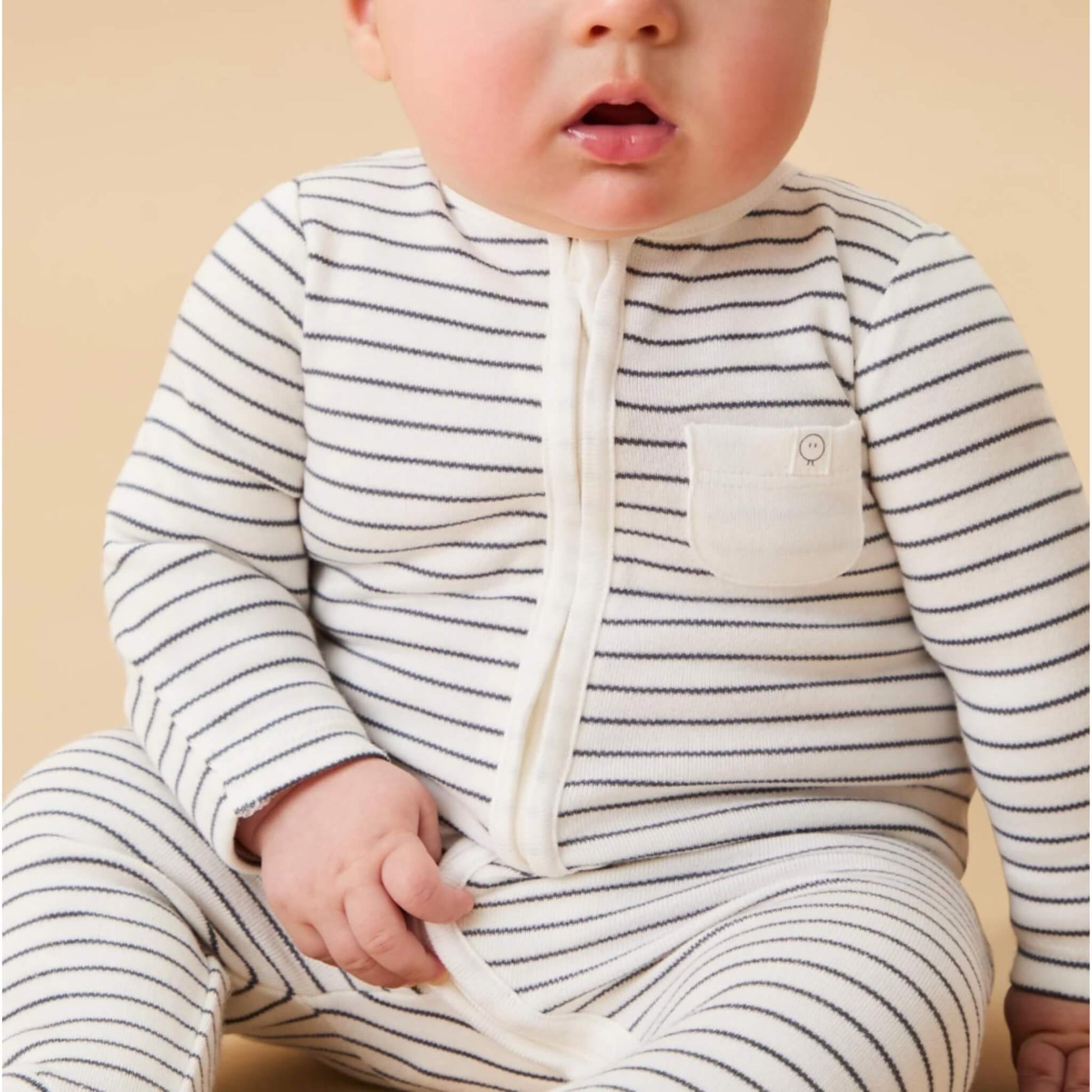 A baby wearing a grey striped sleepsuit, shown in a seated position with the long front zip and small chest pocket visible.