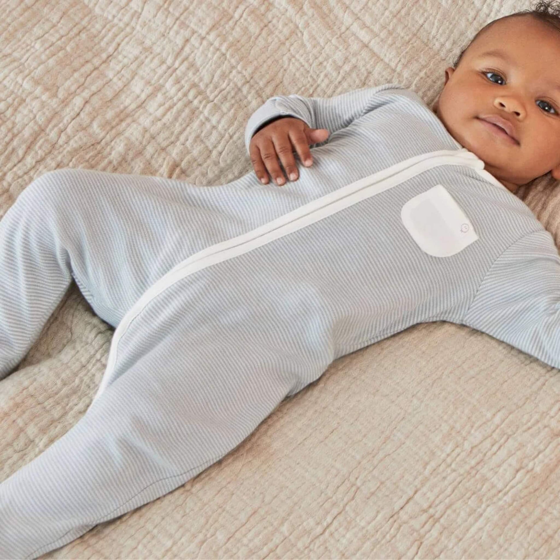 A baby lying on a quilted blanket wearing a blue striped sleepsuit, the full zip visible from neckline to ankle.
