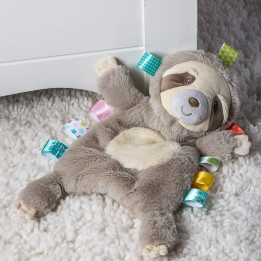 Soft sloth comforter toy with ribbon tags lying on a fluffy white rug beside nursery furniture, showing plush textures and embroidered details.