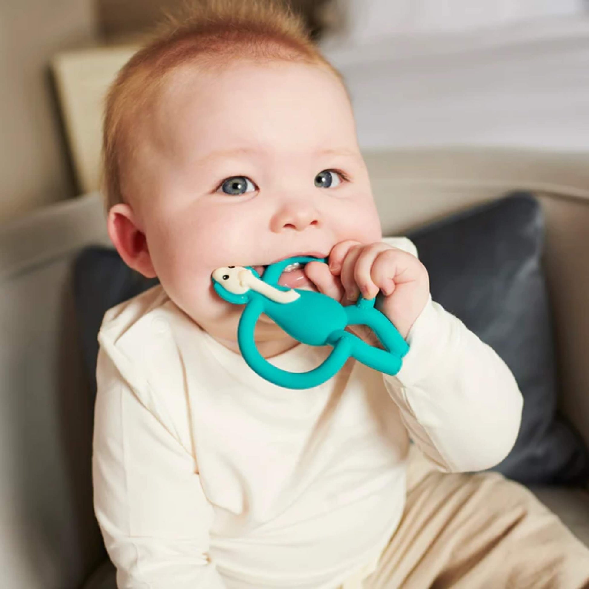 Baby sitting on sofa holding and chewing turquoise elephant silicone teether, demonstrating easy grip handles and soft chewable texture for teething relief.