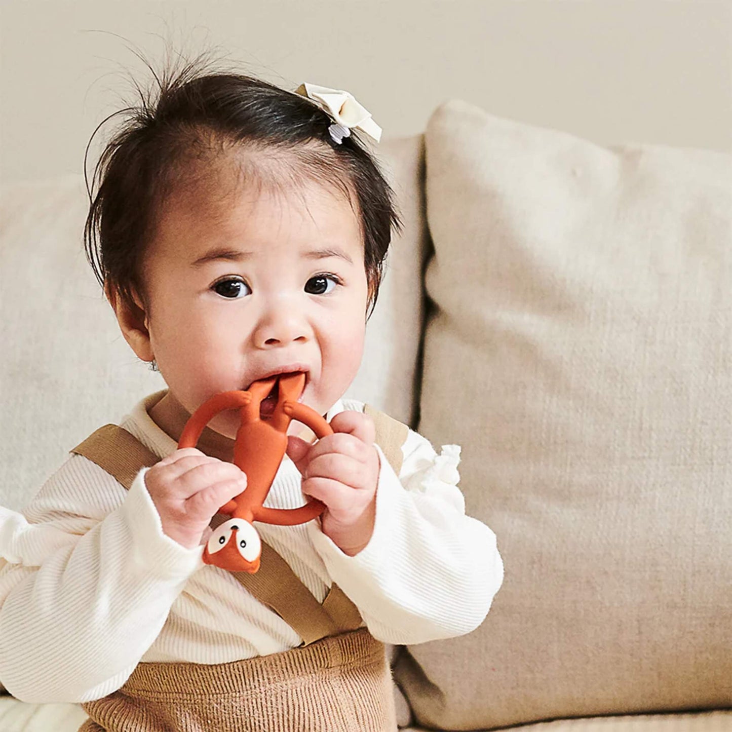Baby holding a fox-shaped teether with both hands while chewing the top section, sitting against a soft sofa cushion.