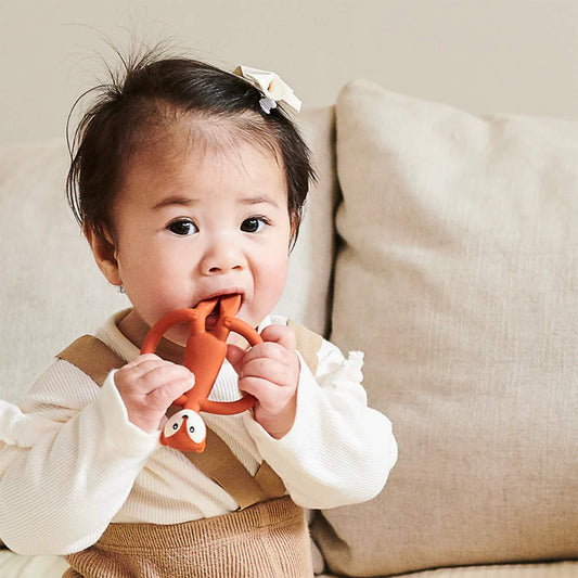 Baby holding a fox-shaped teether with both hands while chewing the top section, sitting against a soft sofa cushion.