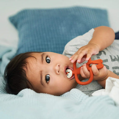 Baby lying on a bed while gripping a fox-shaped teether and chewing the textured head area.