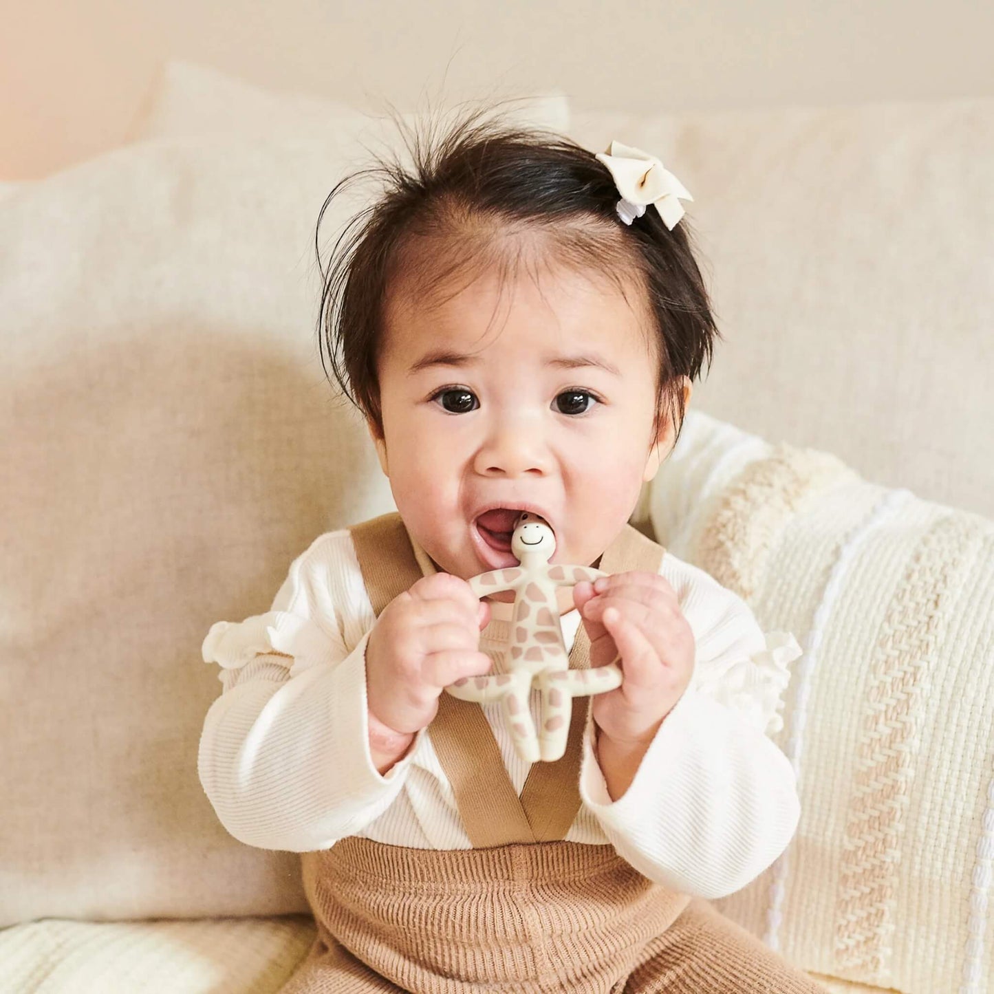 A baby sitting on a soft cushion holding a beige giraffe-shaped silicone teether with both hands while chewing the top section.
