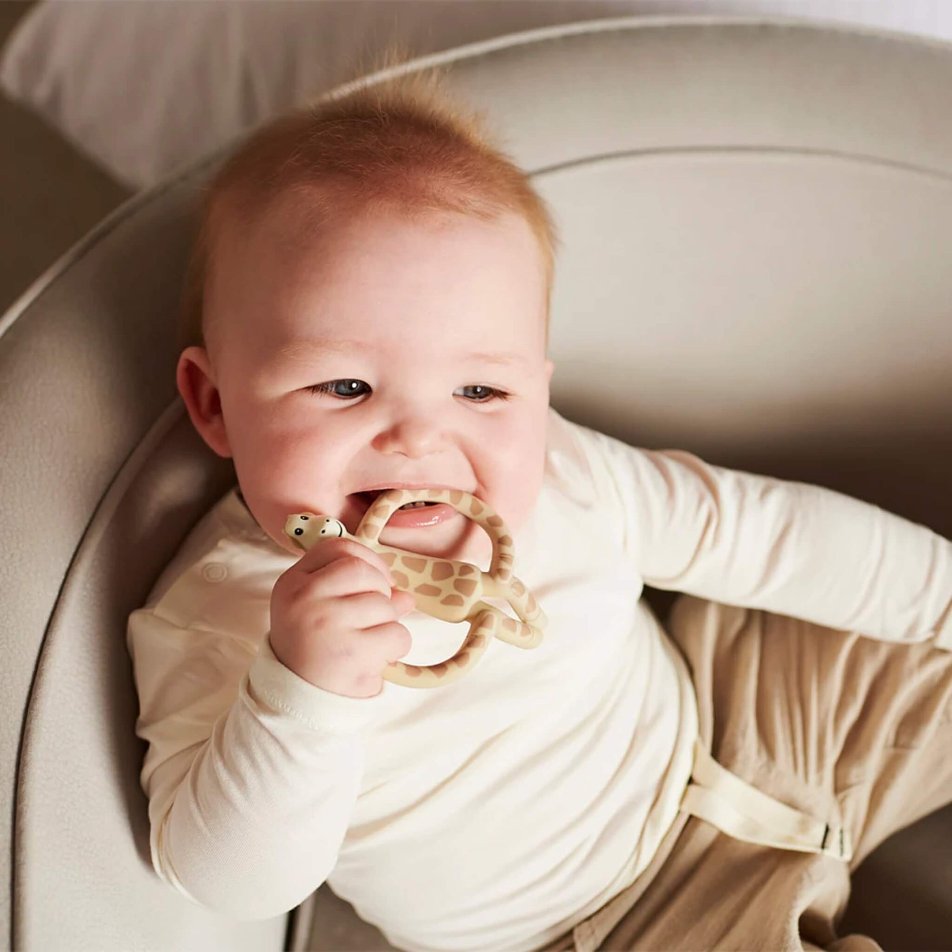 A baby sitting in a cushioned chair holding a giraffe-shaped teether close to their mouth while smiling.