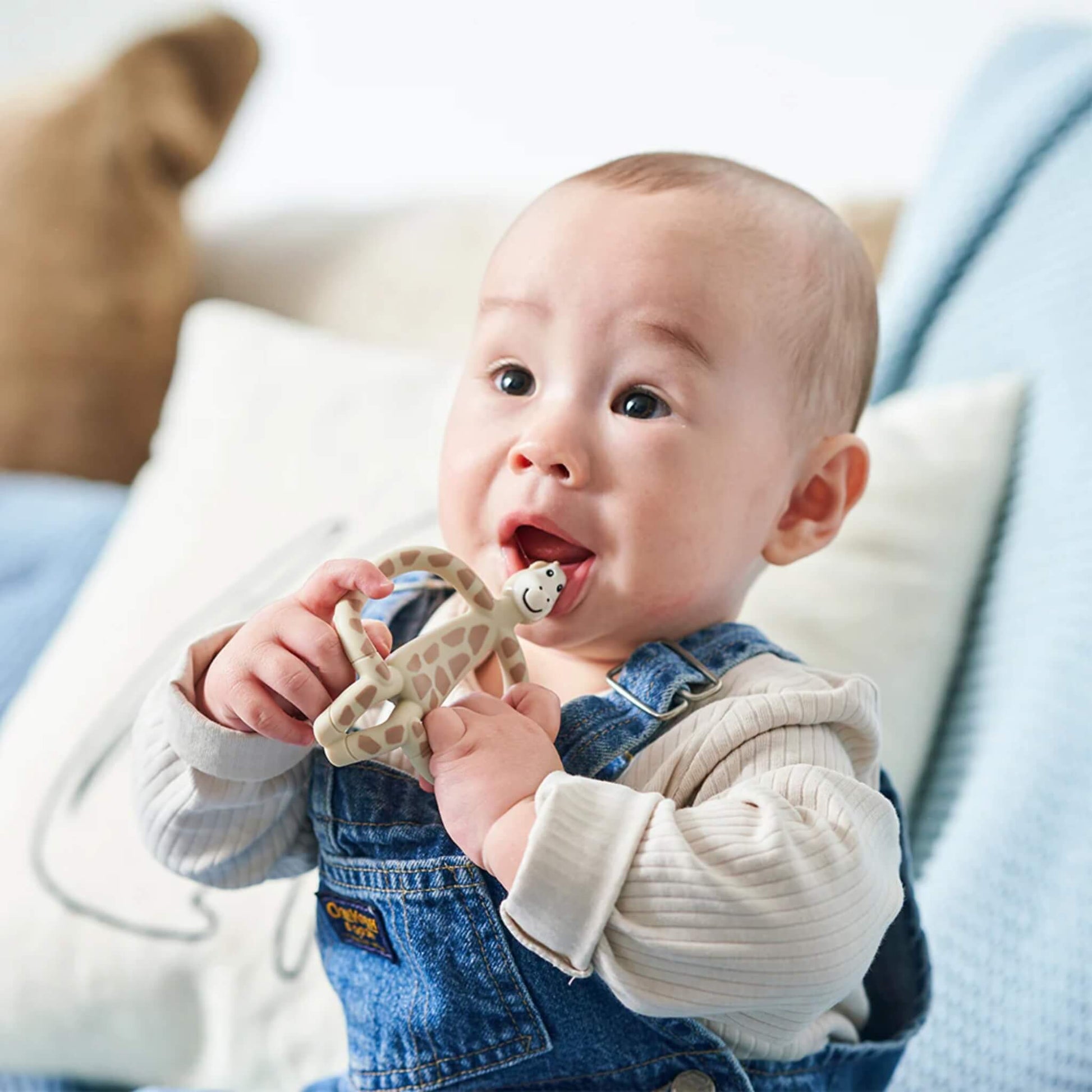 A baby wearing denim overalls holding a giraffe-shaped teether near their mouth while sitting against soft bedding.