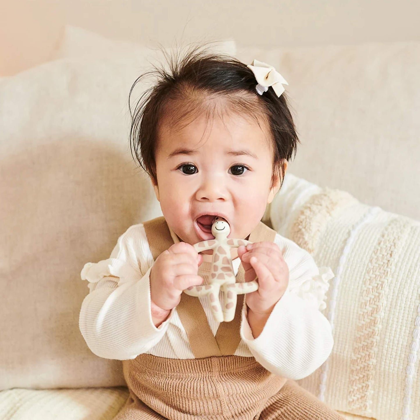 A baby sitting upright on a sofa holding a giraffe-shaped teether with both hands and bringing it toward their mouth.