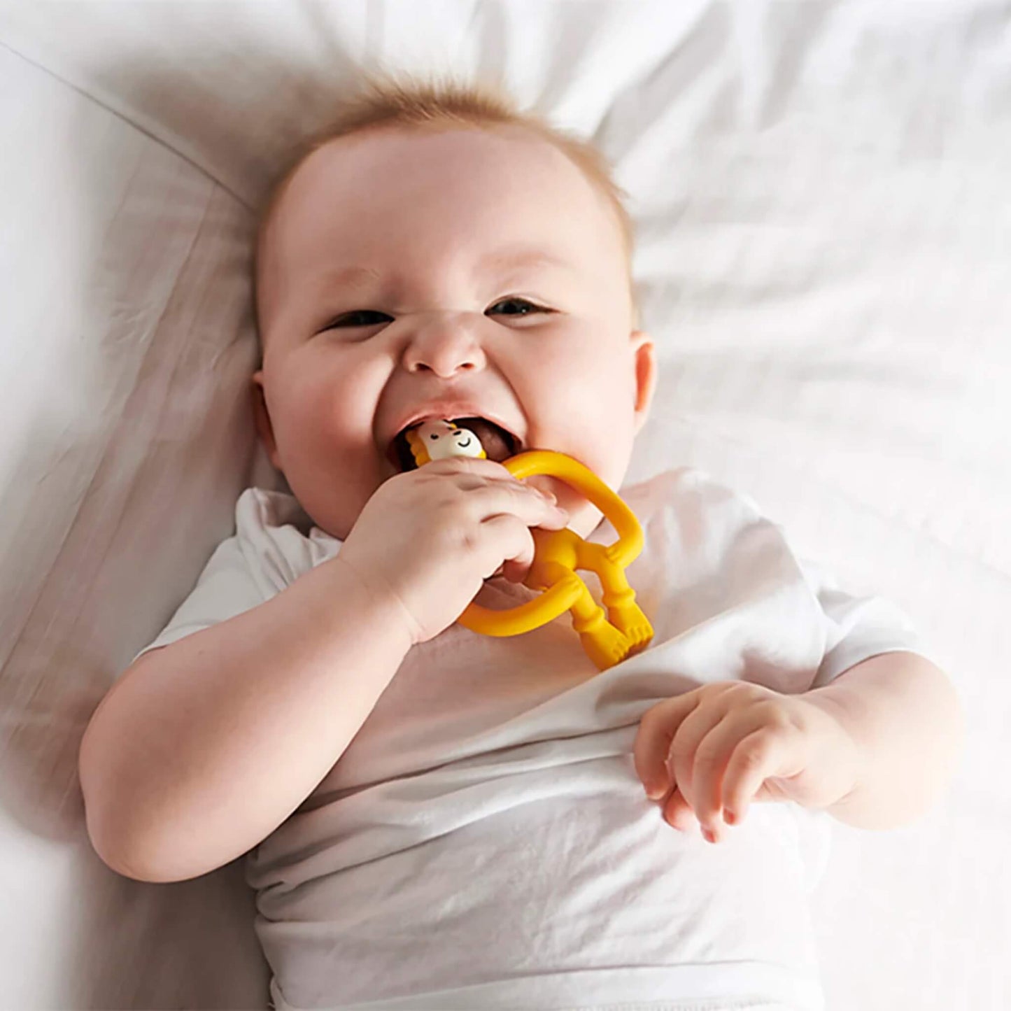 A young baby lying on a soft surface chewing on a yellow lion baby teether.