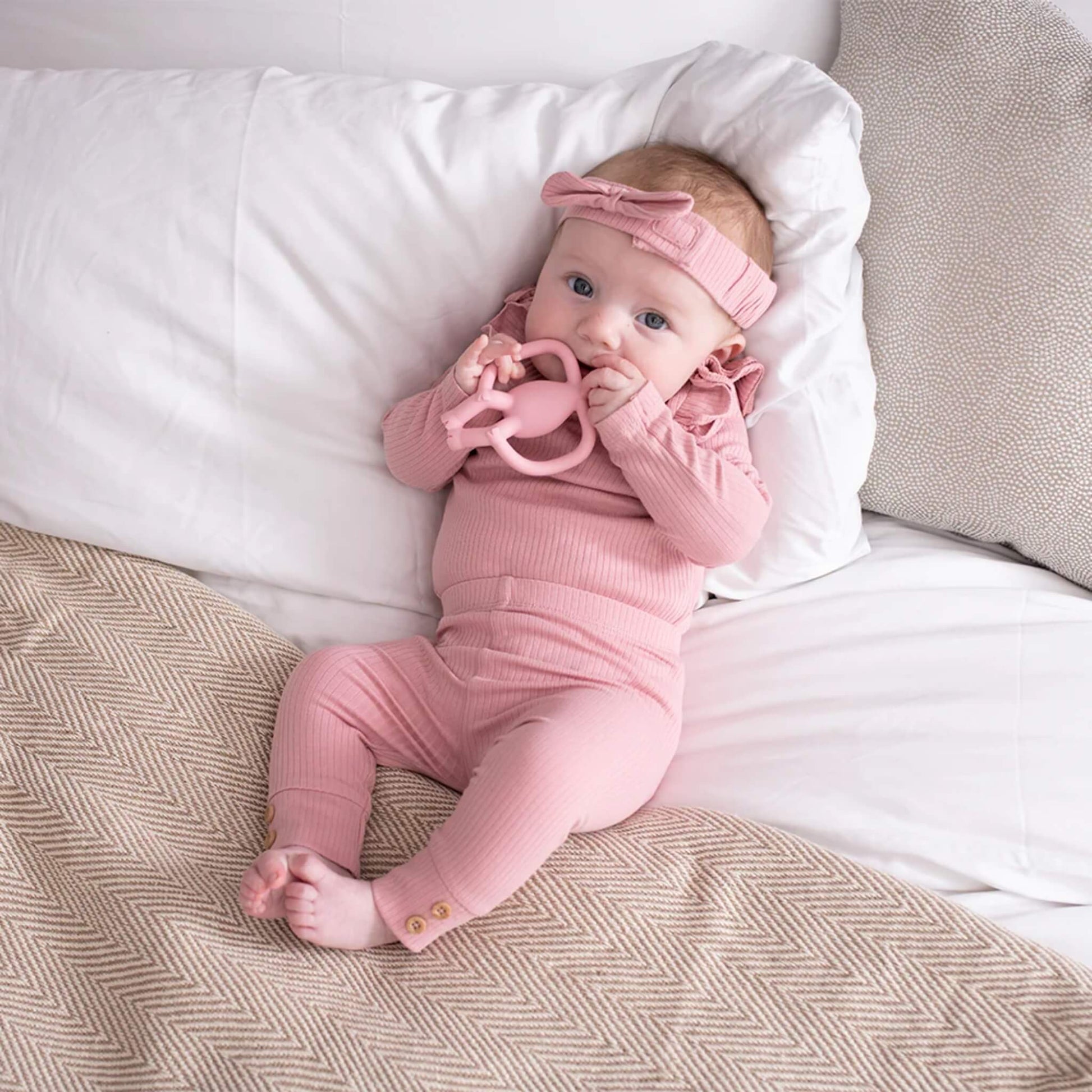A baby lying on a bed while holding a pink animal-shaped teether and bringing it to their mouth.