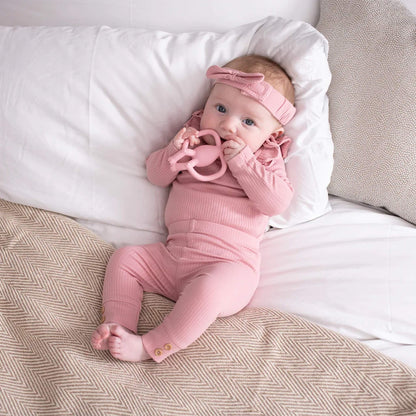 A baby lying on a bed while holding a pink animal-shaped teether and bringing it to their mouth.