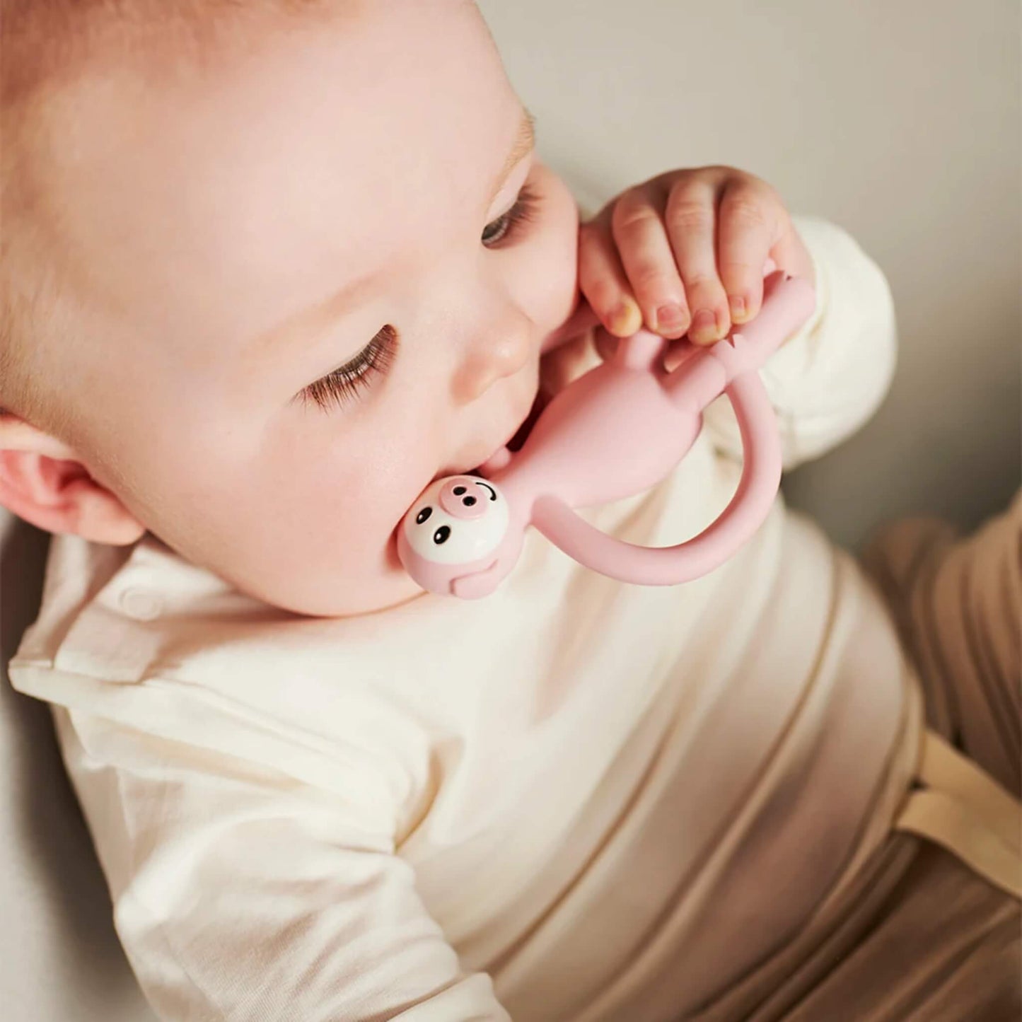A close-up of a baby chewing on a pink silicone teether while gripping it with both hands.