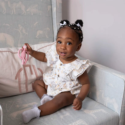A toddler sitting on a small chair and holding up a pink animal-shaped teether with a smile.
