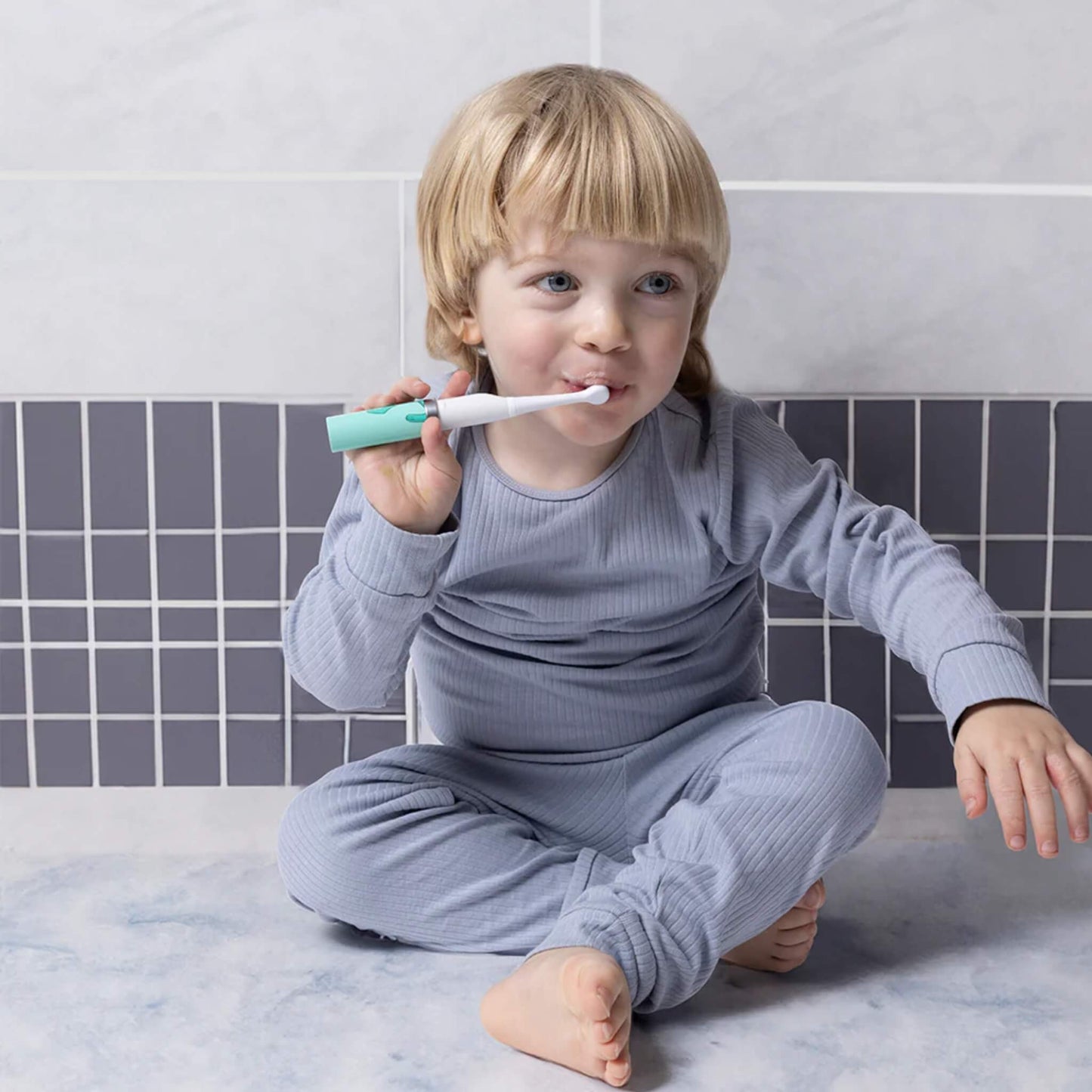 Young child brushing their teeth with a mint-coloured electric toothbrush fitted with one of the replacement heads, seated on a tiled bathroom floor.