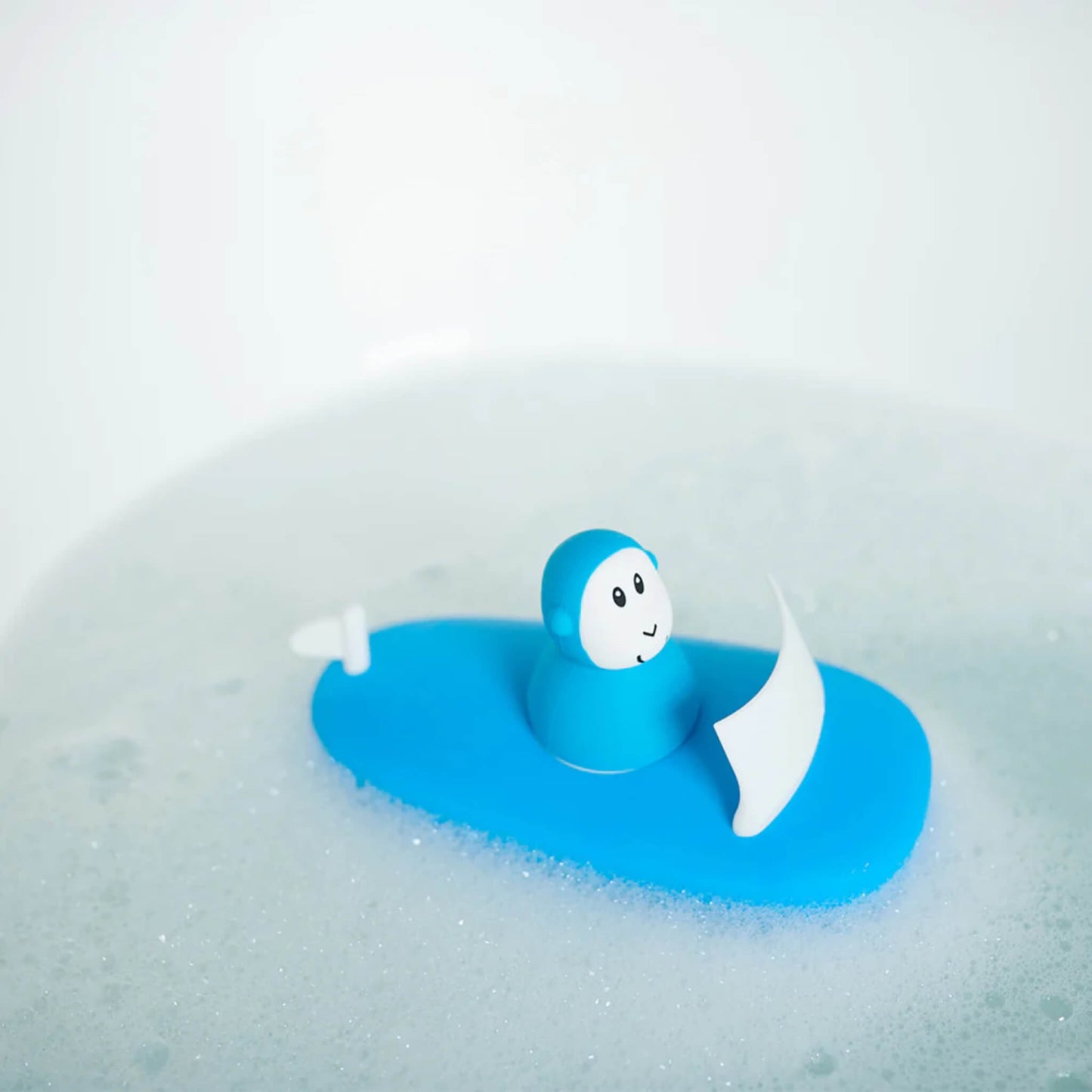 Blue bath toy boat floating among bubbles in a tub, with the monkey figure seated upright on the deck.