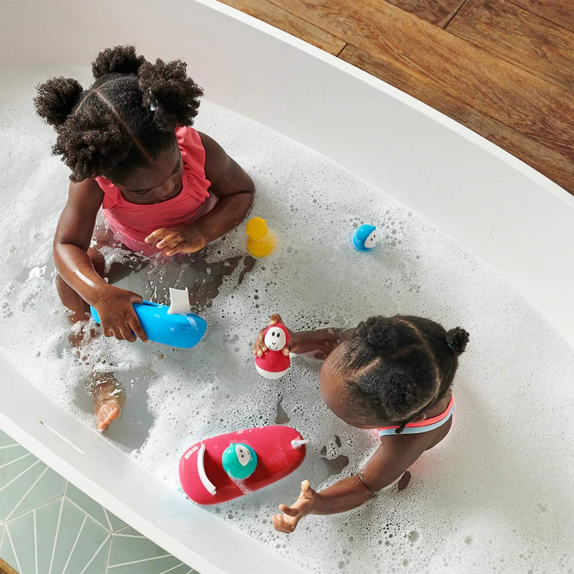 Two young children playing in a bubble-filled bath with several colourful boat toys and matching character figures.
