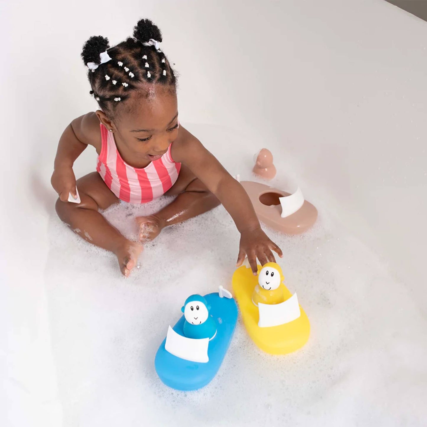 Toddler sitting in a bubble bath playing with blue and yellow silicone boat toys, each with a smiling character figure on top.
