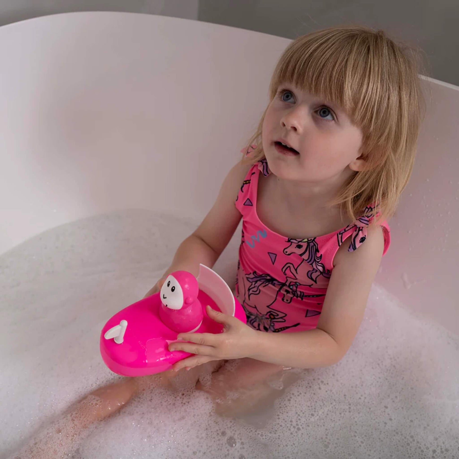 Close-up of a child holding the pink silicone boat toy above bath water, showing the smooth, rounded shape and white sail piece.
