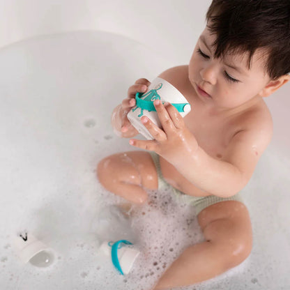 A toddler sitting in a bubble bath holding a small bucket with a character illustration while water drips through the holes at the base.