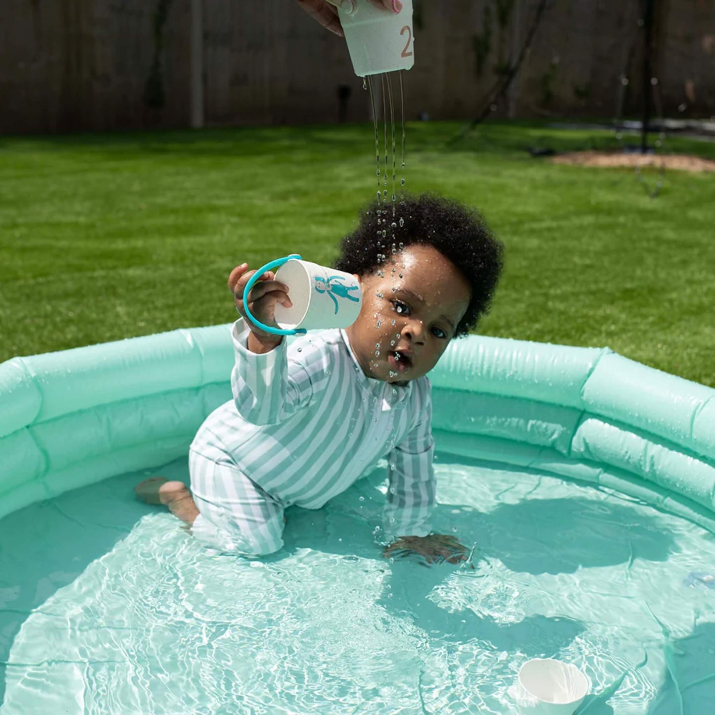 A young child in an outdoor paddling pool holding a small toy bucket while an adult pours water from another bucket above, creating a stream of droplets.