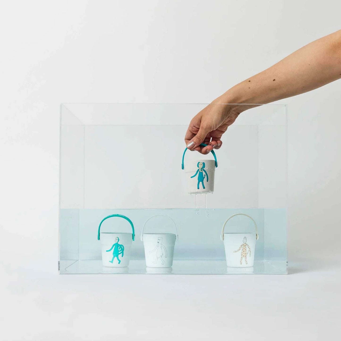 A hand lowering a bucket with a blue handle into a clear water tank while three other buckets sit submerged below the surface.