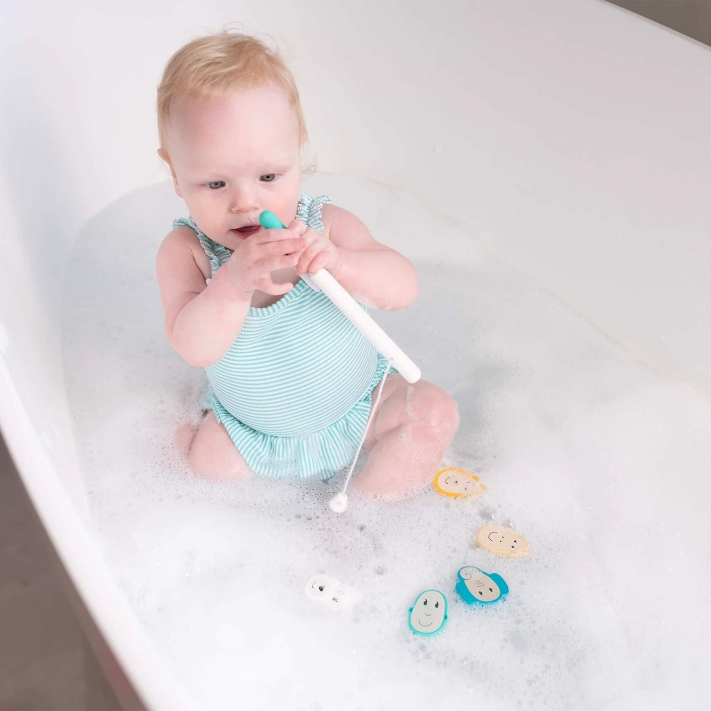 A baby in a bubble bath holding the fishing rod while the animal pieces float nearby.