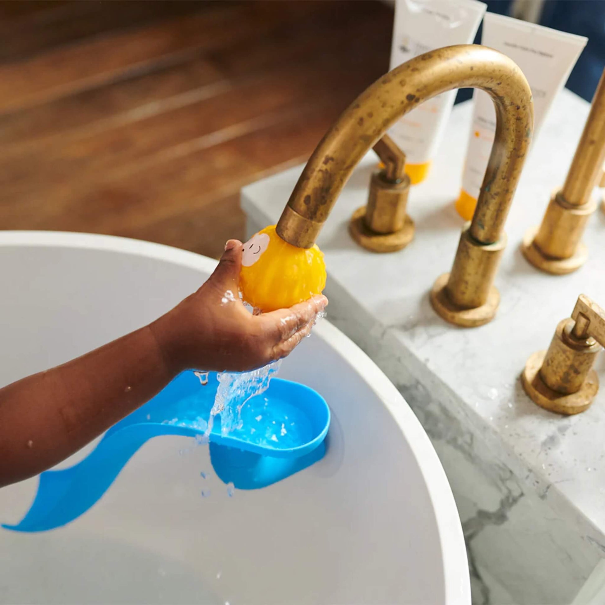 Child’s hand holding a yellow animal character ball under a running tap above a basin, with the blue bath slide resting on the edge below.