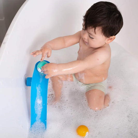 Young child sitting in a bubble-filled bath while placing a turquoise animal ball onto a blue curved slide attached to the bath wall.