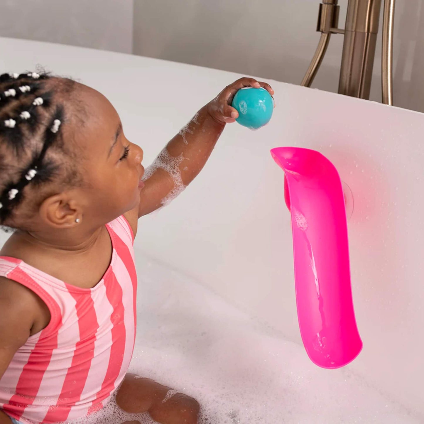 Young child sitting in a bubble-filled bath and holding a turquoise animal ball above a pink curved slide attached to the bath wall.