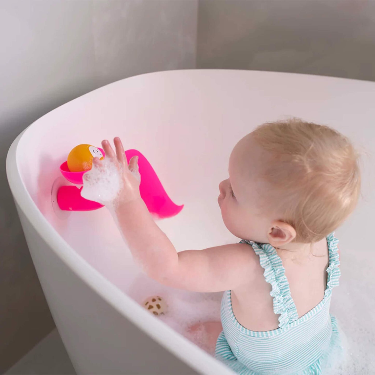 Toddler sitting in a foamy bath while placing a yellow animal ball onto a pink slide attached to the side of the tub.