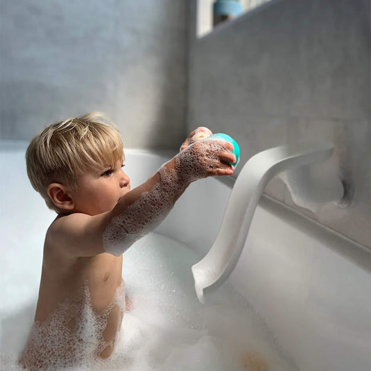 Child in a bubble-filled bath reaching up to place a turquoise animal ball at the top of a white slide fixed to the bath wall.